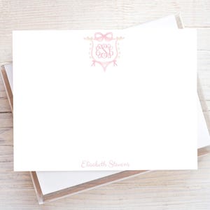 May include: A white rectangular stationery card featuring a light pink decorative crest with a bow at the top and the monogram E S B in the center. The name Elisabeth Stevens is printed in light pink text at the bottom center of the card.