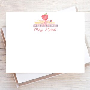 May include: A white rectangular stationery card featuring a watercolor illustration of a stack of books topped with a red apple and a pencil, tied with a pink ribbon. The text "Mrs. Hood" is printed in a red script font below the illustration.