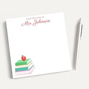 May include: A square white notepad featuring the red printed text FROM THE DESK OF Mrs. Johnson at the top. The bottom left corner displays a stack of four books in green, yellow, teal, and pink with a red apple on top. A silver pen lies nearby.