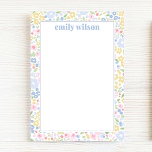 May include: A floral patterned notepad with the name "emily wilson" in light blue. The notepad is surrounded by a border of small flowers in pastel colors, including pink, yellow, and blue. A wooden pencil rests on the right side.