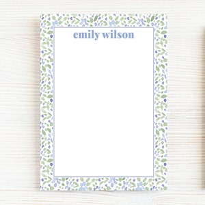 May include: A personalized notepad with a blue border and a floral pattern in shades of blue and green. The name "emily wilson" is printed at the top. A wooden pencil rests on the side, suggesting a stationery theme.
