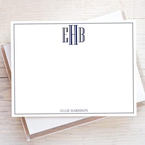 May include: White stationery cards with a navy blue border. The cards feature a navy blue monogram with the initials EHB at the top and the name ELLIE HARRISON at the bottom. The cards are stacked in a clear plastic box.