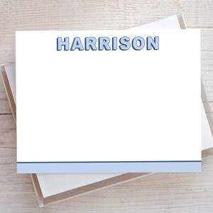 May include: White stationery featuring the name "HARRISON" in blue block letters at the top. A blue horizontal stripe is at the bottom. The stationery is in a clear plastic box, on a light wood surface.