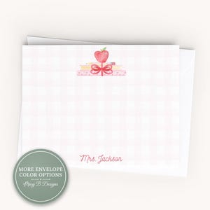 May include: A white stationery card with a light pink gingham pattern, featuring a watercolor illustration of a red apple, a yellow pencil, and a pink bow on a stack of books. The text Mrs. Jackson is printed in pink script at the bottom center.