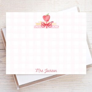 May include: A rectangular white stationery card featuring a faint pink checkered pattern. The top center displays a watercolor illustration of a red apple, a pencil, and a stack of books tied with a red ribbon. The name Mrs. Jackson is printed in red.