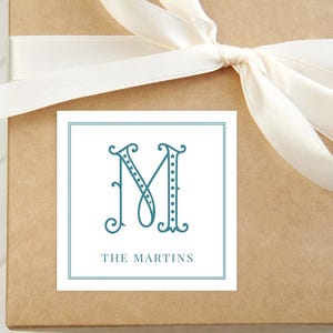 May include: A square gift tag with a teal letter "M" in an elegant, ornate font, with the name "THE MARTINS" below. The tag is on a brown gift box with a cream-colored ribbon tied in a bow.
