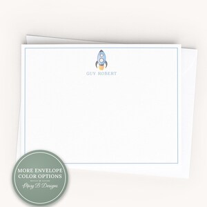 May include: A white rectangular stationery card with a thin blue border, featuring a small watercolor illustration of a blue rocket ship and the printed name GUY ROBERT. A circular sage green badge displays the text MORE ENVELOPE COLOR OPTIONS.