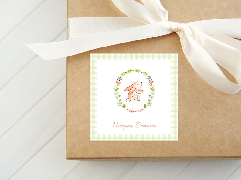 May include: A square gift tag with a light green gingham border features a watercolor illustration of a bunny inside a floral wreath. The name "Harper Brown" is printed in a soft pink font below the bunny. The tag is tied to a brown box with a cream-colored ribbon.