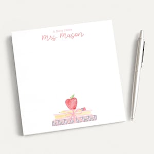 May include: A square white notepad with the text A Note From Mrs. Mason printed in pink at the top. The bottom features a watercolor illustration of a stack of books, a yellow pencil, and a red apple tied with a pink ribbon. A silver pen sits nearby.
