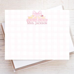 May include: A rectangular stationery card featuring a light pink plaid pattern background. At the top center, a watercolor illustration shows a stack of books topped with a pink bow and a pencil. The text MRS. JACKSON is printed in pink.