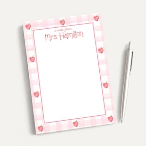 May include: A rectangular notepad featuring a light pink and white gingham border decorated with small red apple illustrations. The center is white with a pink border and the text "a note from Mrs. Hamilton" printed in red. A silver ballpoint pen.
