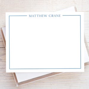 May include: White stationery with a navy blue border and the name "MATTHEW CRANE" printed at the top. The stationery is stacked in a clear plastic box, resting on a light wood surface.