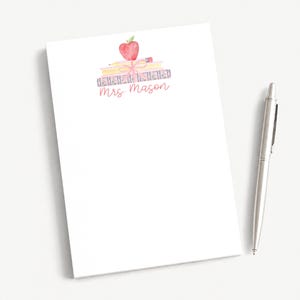 May include: A white rectangular notepad featuring a watercolor illustration of a stack of books topped with a red apple and a pencil, tied with a pink ribbon. The text Mrs Mason is printed in a script font below the illustration, alongside a silver pen.