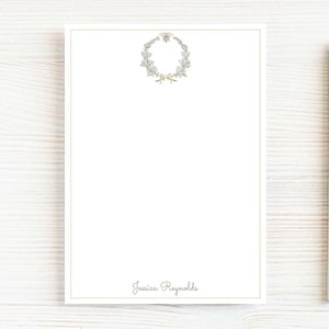 May include: A rectangular white stationery sheet featuring a thin gold border, a central top wreath illustration with a small bee, and the name Jessica Reynolds printed in a grey script font at the bottom, accompanied by a wooden pencil.