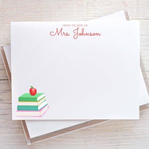 May include: A white rectangular stationery note card featuring the red printed text FROM THE DESK OF Mrs. Johnson at the top center. The bottom left corner displays a stack of four colorful books topped with a single bright red apple illustration.