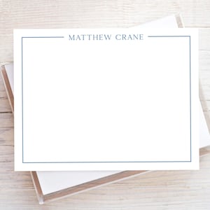 May include: A rectangular white stationery card featuring a thin blue border and the name MATTHEW CRANE printed in blue capital letters at the top center. The card is stacked on top of other white cards within a clear plastic holder.