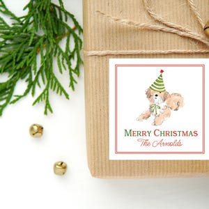 May include: A square white sticker with a red border featuring a watercolor illustration of a light brown dog wearing a green and white striped party hat and a green bow. The text reads Merry Christmas and The Arnolds in red and green script.