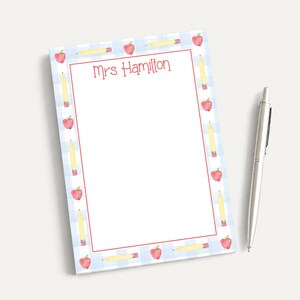 May include: A rectangular notepad with a light blue and white gingham pattern border featuring watercolor illustrations of red apples and yellow pencils. The text Mrs. Hamilton is printed in red at the top. A silver metal pen lies to the right.