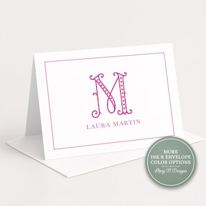 Op de afbeelding: Een witte gevouwen kaart met een roze rand en een groot, gestileerd roze letter M monogram. Onder het monogram staat de naam LAURA MARTIN in het roze gedrukt. Een ronde saliegroene badge met witte tekst vermeldt MORE INK & ENVELOPE COLOR OPTIONS.