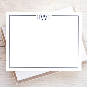 May include: White stationery with a navy blue border and initials "HWH" at the top. The stationery is stacked on top of other stationery in a clear plastic box. The background is a light wood surface.
