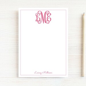 May include: A white rectangular notepad with a thin pink border, featuring a pink decorative monogram with the letters LME at the top center and the name Lainey Mathison printed in pink script at the bottom, accompanied by a wooden pencil.