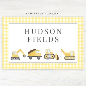 May include: Laminated placemat with a yellow gingham border. The center features the text "HUDSON FIELDS" above illustrations of yellow construction vehicles: an excavator, dump truck, bulldozer, and crane.