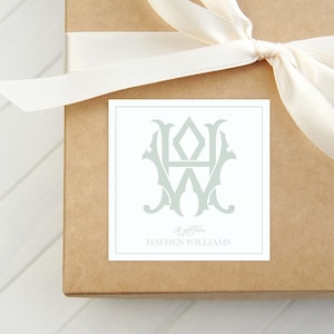 May include: A gift box wrapped in brown paper with a cream-colored ribbon tied in a bow. A square label with a monogram in a light green color and the text "Hayden Williams" is centered on the box.