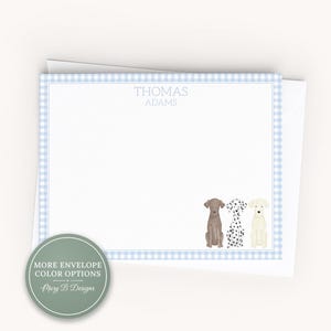 May include: A white rectangular stationery card featuring a light blue gingham border. The text THOMAS ADAMS is printed in light blue at the top center. Three illustrated dogs, one brown, one spotted, and one cream, sit at the bottom right corner.