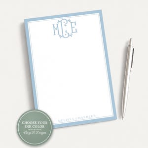 May include: A rectangular notepad with a light blue border, featuring a centered light blue monogram with the letters M, C, and E at the top and the name MELISSA CHANDLER printed at the bottom. A silver ballpoint pen rests beside the notepad.