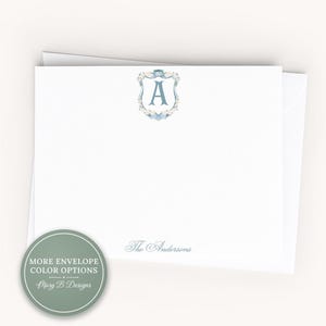 May include: A white stationery card featuring a centered blue letter A enclosed in a floral crest design. The text The Andersons is printed in blue at the bottom. A circular green badge in the corner displays the text MORE ENVELOPE COLOR OPTIONS.