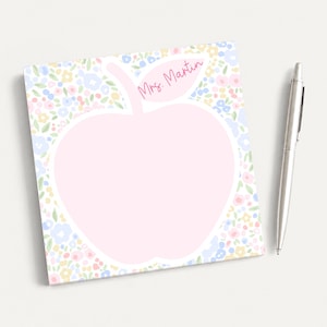 May include: A square notepad featuring a large, central apple-shaped area in light pink for writing, surrounded by a colorful floral pattern of small blue, yellow, and pink flowers. The text Mrs. Martin is printed in pink on the apple leaf.