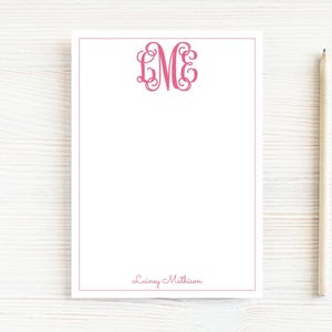 May include: A white notepad with a pink border and a pink monogram in the upper center. The monogram features the letters L, M, and E. The name "Lainey Mathison" is written in pink script at the bottom. A light-colored pencil rests on the right side.