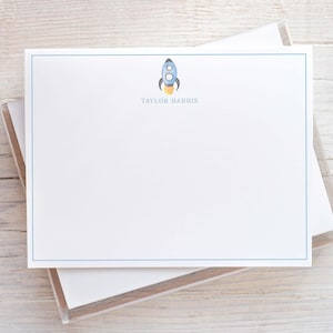 May include: A white rectangular stationery card featuring a thin blue border, a small blue and yellow rocket ship illustration at the top center, and the name TAYLOR HARRIS printed in blue capital letters directly below the rocket graphic.