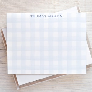 May include: A stack of personalized stationery featuring a light blue and white gingham pattern. The name "THOMAS MARTIN" is printed in a dark blue font at the top of the stationery. The stationery is displayed in a clear plastic box.