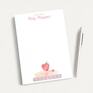 May include: A white rectangular notepad with the printed text A Note From Mrs Mason in pink script. The bottom features a watercolor illustration of a stack of books tied with a pink ribbon, topped with a red apple and a yellow pencil.