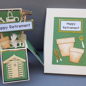 Op de afbeelding: Een groene en gouden pop-upkaart met een tuinmotief. De kaart toont een klein schuurtje, tuinhandschoenen, een gieter, een hark, een schep en twee bloempotten. De kaart luidt "Happy Retirement".