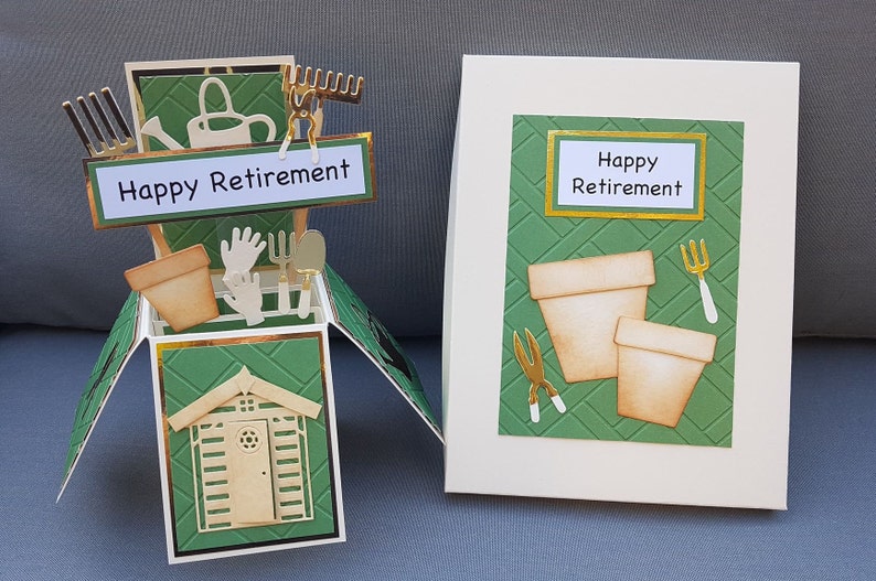 Op de afbeelding: Een groene en gouden pop-upkaart met een tuinmotief. De kaart toont een klein schuurtje, tuinhandschoenen, een gieter, een hark, een schep en twee bloempotten. De kaart luidt "Happy Retirement".