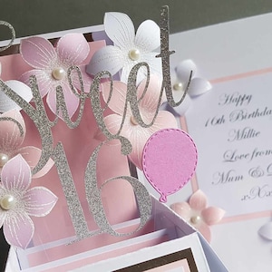 May include: A pink and white 3D pop-up card with the words "Sweet 16" in silver glitter. The card is decorated with pink and white flowers and a pink balloon. The card also includes the text "Happy 16th Birthday Millie Love from Mum & Dad xx".