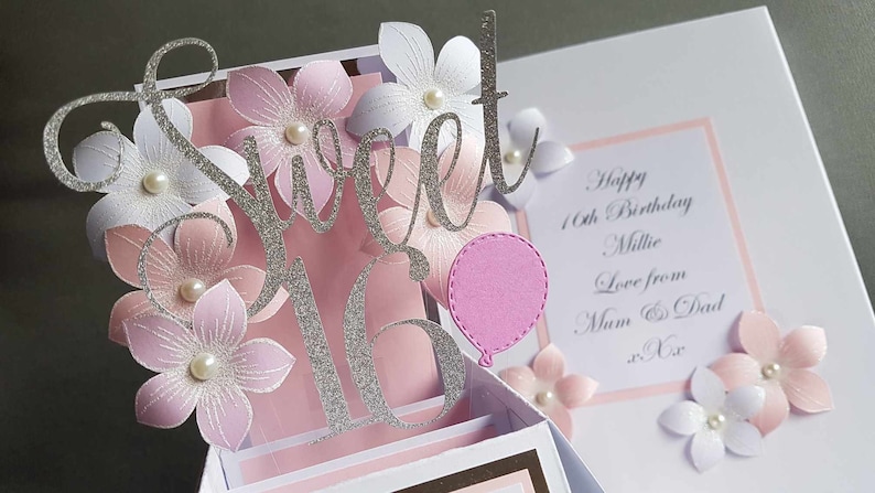 May include: A pink and white 3D pop-up card with the words "Sweet 16" in silver glitter. The card is decorated with pink and white flowers and a pink balloon. The card also includes the text "Happy 16th Birthday Millie Love from Mum & Dad xx".