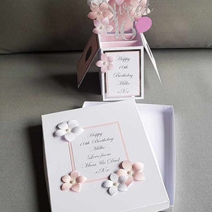 May include: A white box with a pink and white floral design. The box is open and the inside is white. The box has a message that says "Happy 16th Birthday Millie Love from Mum & Dad".