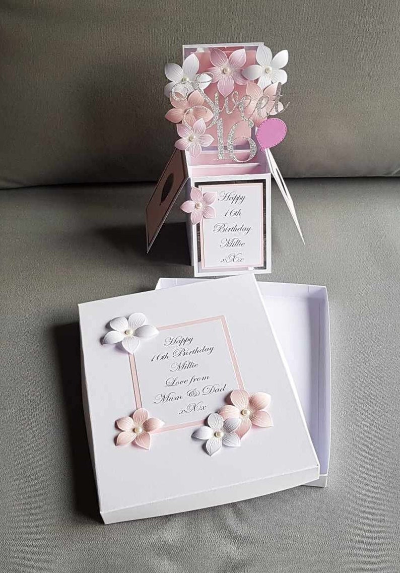 May include: A white box with a pink and white floral design. The box is open and the inside is white. The box has a message that says "Happy 16th Birthday Millie Love from Mum & Dad".