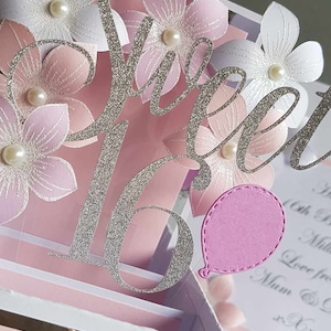 May include: A pink and white 3D pop-up card with a silver glitter 'Sweet 16' and a pink balloon. The card has a white background with a gold border and the words 'Happy Birthday' written in black.