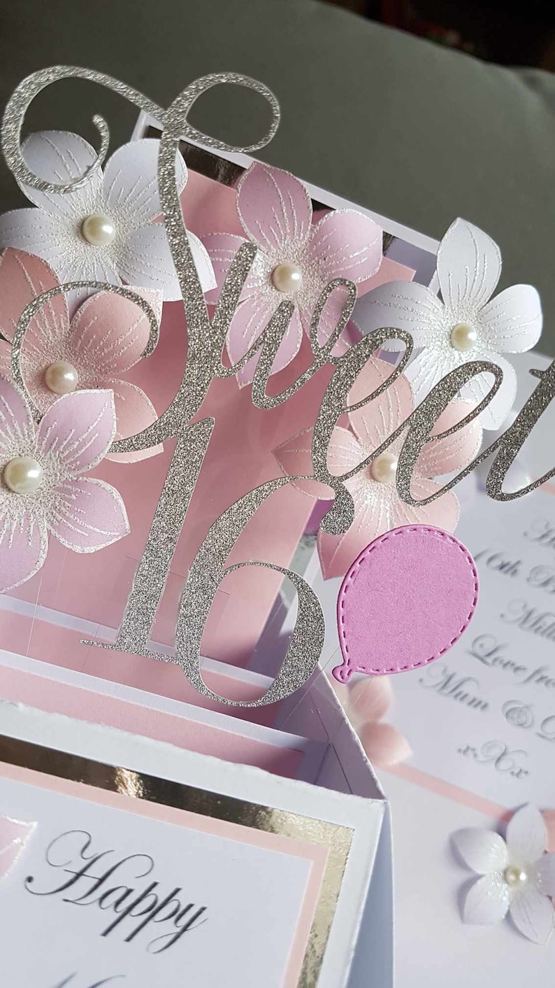 May include: A pink and white 3D pop-up card with a silver glitter 'Sweet 16' and a pink balloon. The card has a white background with a gold border and the words 'Happy Birthday' written in black.