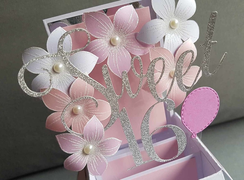 May include: A silver glitter card with the words "Sweet 16" and pink flowers. The card is decorated with white pearls and a pink balloon.