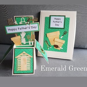 Op de afbeelding: Een groene pop-upkaart met een schuur, tuingereedschap en bloempotten. De kaart heeft de tekst "Happy Father's Day". De kaart ligt op een grijs oppervlak met de woorden "Emerald Green" in wit.