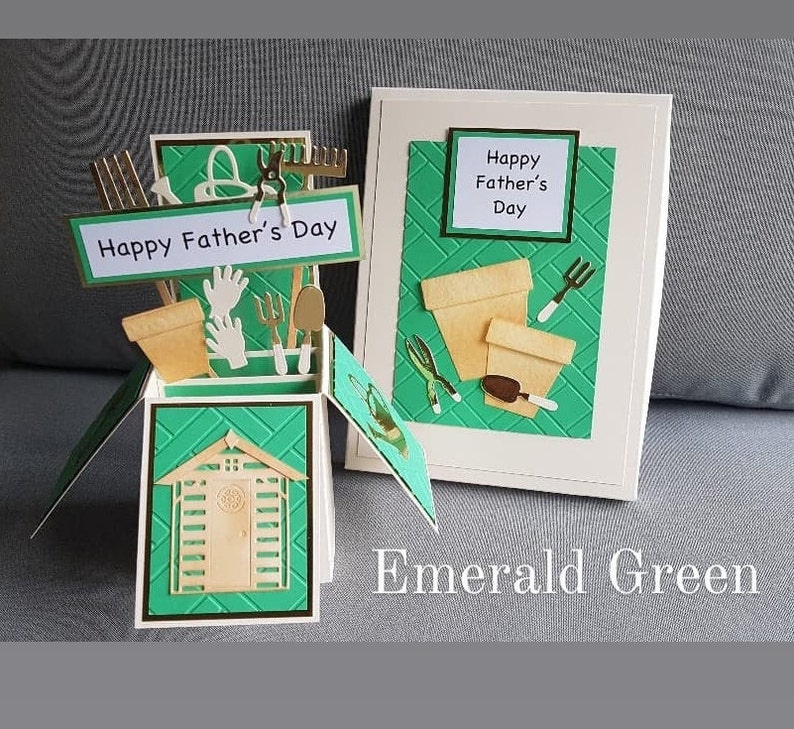Op de afbeelding: Een groene pop-upkaart met een schuur, tuingereedschap en bloempotten. De kaart heeft de tekst "Happy Father's Day". De kaart ligt op een grijs oppervlak met de woorden "Emerald Green" in wit.