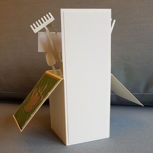Op de afbeelding: Een witte kaart met een pop-up ontwerp met tuingereedschap, waaronder een hark, een schop en een gieter. De kaart is gevouwen en staat rechtop op een grijs oppervlak.