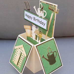 Op de afbeelding: Een groene en gouden pop-upkaart met een tuinmotief. De kaart toont een klein schuurtje, tuingereedschap en een gieter. De kaart luidt "Happy Birthday".