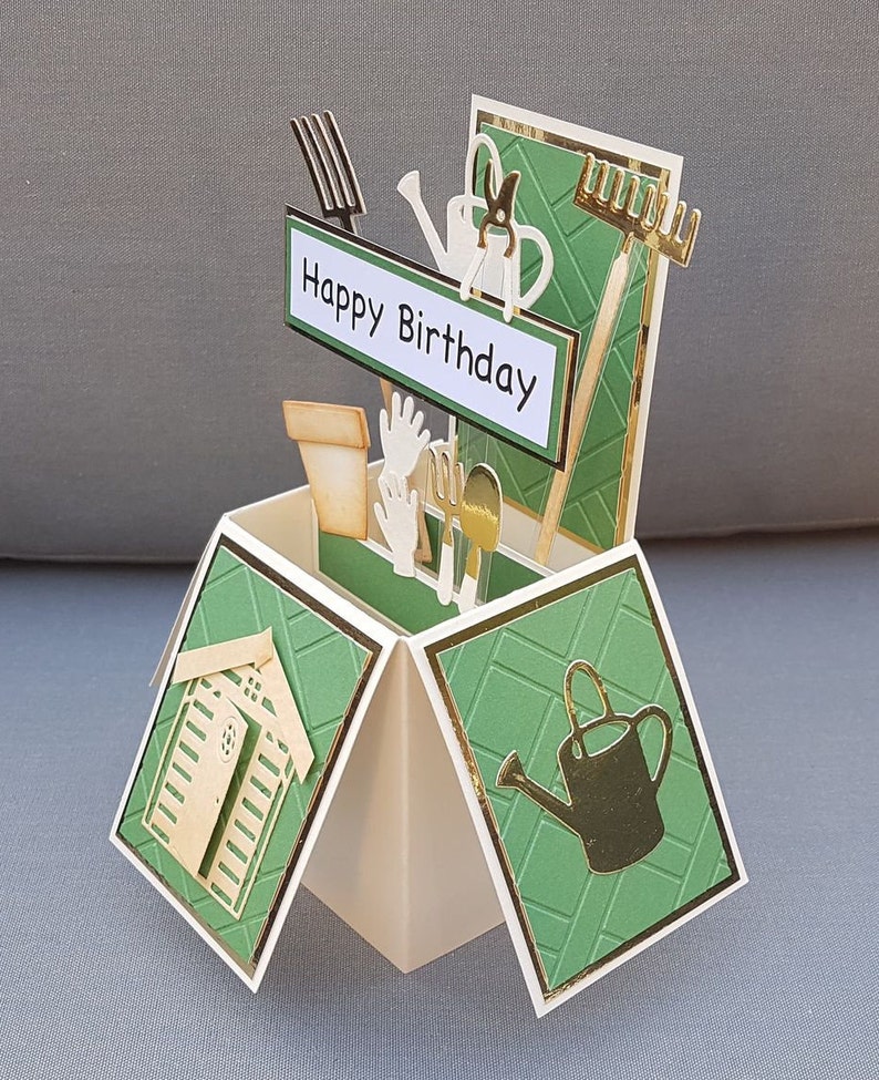 Op de afbeelding: Een groene en gouden pop-upkaart met een tuinmotief. De kaart toont een klein schuurtje, tuingereedschap en een gieter. De kaart luidt "Happy Birthday".