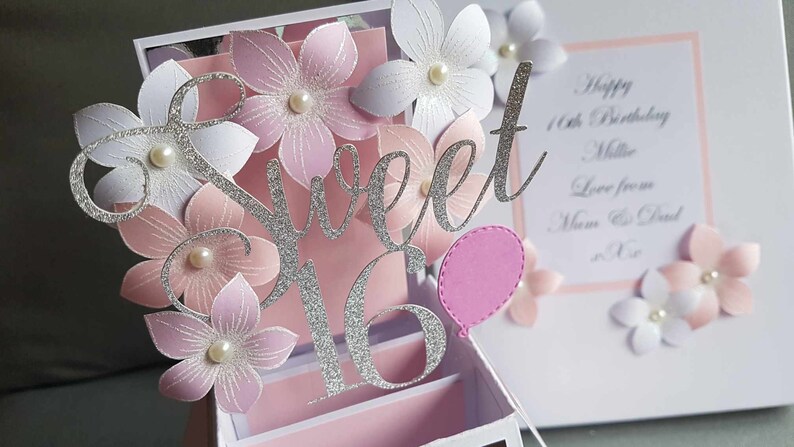 May include: A silver glitter 'Sweet 16' pop-up card with pink and white paper flowers and a pink balloon. The card has a white background with a pink border and the text 'Happy 16th Birthday Millie Love from Mum & Dad xxx'.